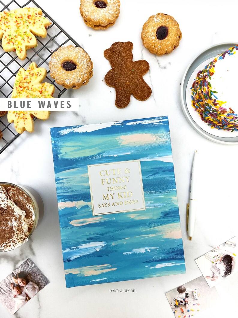 Puede incluir: Un libro azul y blanco con el t&iacute;tulo "Cute & Funny Things My Kid Says and Does" de Daisy & Decor. El libro est&aacute; rodeado de varios art&iacute;culos, que incluyen galletas, un oso de peluche y una taza de caf&eacute;.