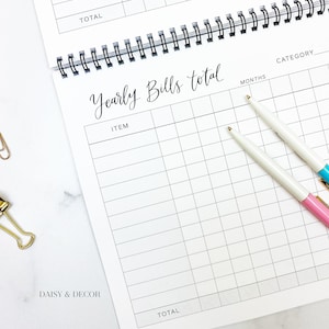 K&ouml;nnte beinhalten: Ein wei&szlig;er Spiralplaner, der auf einer Seite "Yearly Bills total" ge&ouml;ffnet ist. Die Seite hat ein Rasterlayout f&uuml;r detaillierte Ausgaben. Zwei Stifte mit wei&szlig;en Sch&auml;ften und goldenen Spitzen, einer mit rosa Ende und der andere mit blauem Ende, liegen auf der Seite.