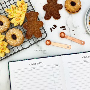 May include: A white cookbook with the words "CONTENTS" and "RECIPE" printed on the cover. The cookbook is open to a page with lined paper and the words "PAGE #" printed at the top. The cookbook is lying on a white surface with a black wire rack holding various cookies and measuring spoons.