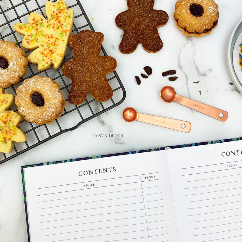 May include: A white cookbook with the words "CONTENTS" and "RECIPE" printed on the cover. The cookbook is open to a page with lined paper and the words "PAGE #" printed at the top. The cookbook is lying on a white surface with a black wire rack holding various cookies and measuring spoons.