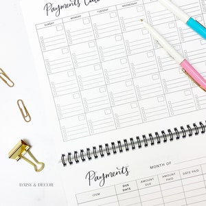 K&ouml;nnte beinhalten: Ein wei&szlig;er, spiralgebundener Zahlungskalender mit schwarzem Raster. Der Kalender enth&auml;lt den Text "Payments Calendar" in eleganter Schrift. Zwei Stifte, einer mit rosa und einer mit blauem Akzent, sowie goldene B&uuml;roklammern sind ebenfalls sichtbar.