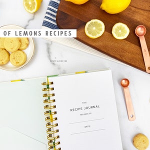 Op de afbeelding: Een wit receptenboek met een gouden spiraalbinding en de tekst "This Recipe Journal Belongs To" en "Date:" op de kaft. Het boek is open op een lege pagina. Er liggen citroenen, citroenschijfjes, maatlepels en citroenkoekjes op een wit marmeren oppervlak.