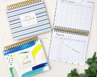 May include: Three spiral-bound bill tracker journals with gold coils. One journal has blue and white stripes, another has a watercolor design, and the third is open, displaying a payment calendar and payment log. A white pen rests on the open journal.