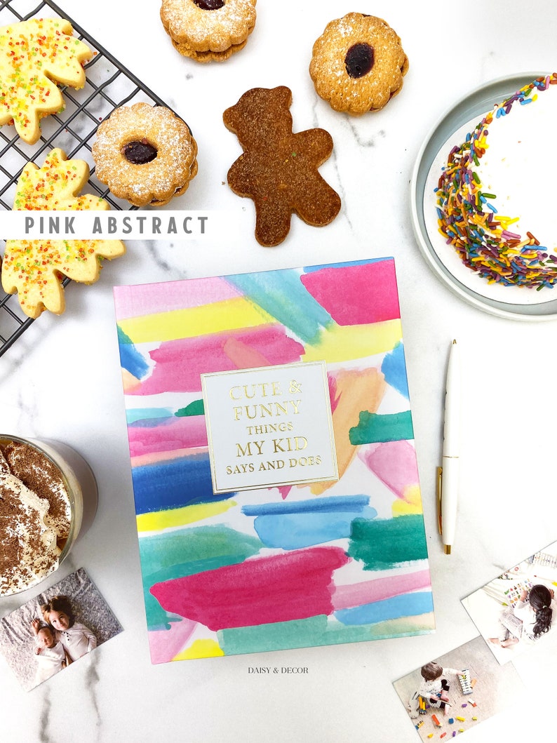 Puede incluir: Un diario de acuarela rosa, azul y amarillo con el texto "Cute & Funny Things My Kid Says and Does" y "Daisy & Decor" en la portada. El diario est&aacute; rodeado de galletas, un pastel y fotos.