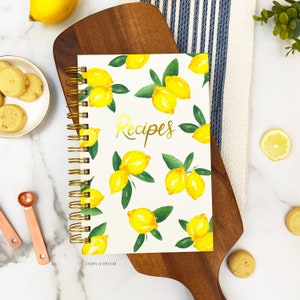Op de afbeelding: Een wit spiraalgebonden receptenboek met een citroenmotief en het woord "Recepten" in gouden letters. Het boek ligt op een houten snijplank met een citroen en koekjes op de achtergrond.