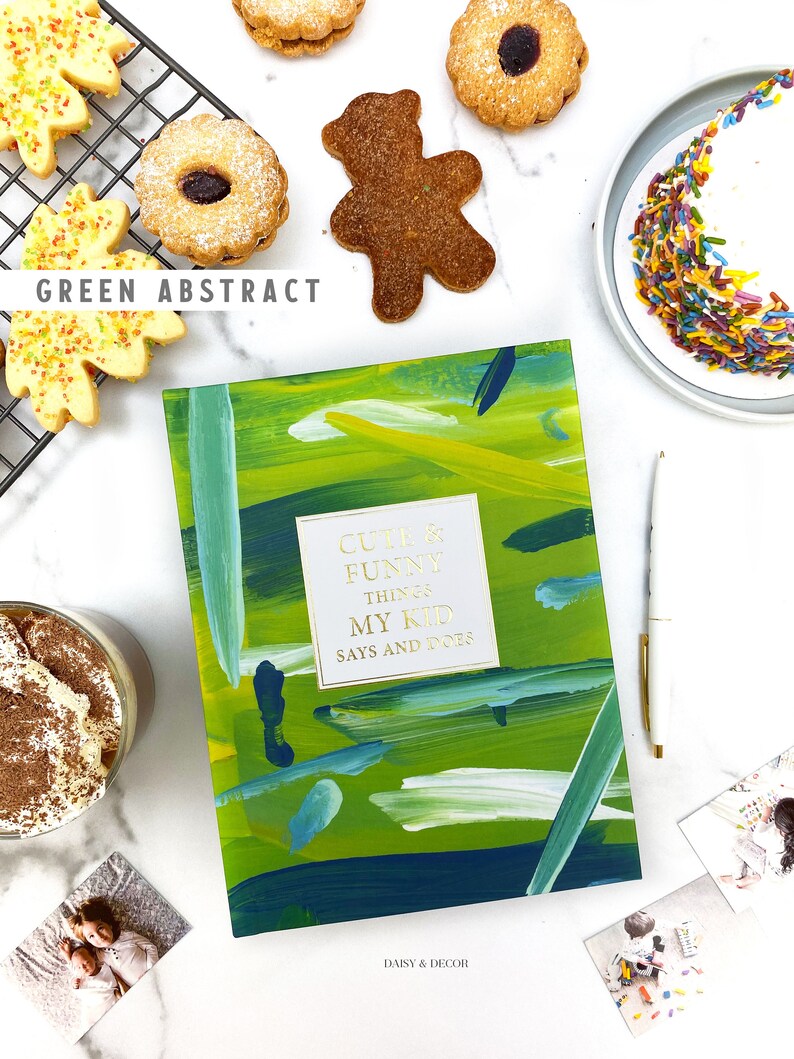 Puede incluir: Un diario verde con pinceladas abstractas y el texto "Cute & Funny Things My Kid Says and Does" en la portada. El diario est&aacute; abierto para revelar una p&aacute;gina en blanco. El diario est&aacute; rodeado de varios art&iacute;culos, que incluyen galletas, un cupcake, una taza de caf&eacute; y fotos.