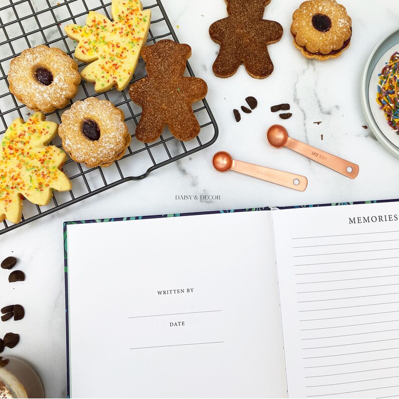 May include: A white notebook with a blue and green floral design on the cover. The notebook is open to a blank page with the words "Written By" and "Date" printed at the top. The notebook is surrounded by various cookies, including gingerbread men, leaf-shaped cookies, and round cookies with sprinkles. There are also two small copper measuring spoons on the table.