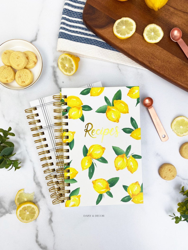 Op de afbeelding: Een wit spiraalgebonden receptenboek met een citroenpatroon en gouden letters die "Recepten" zeggen. Het boek ligt op een wit marmeren oppervlak met citroenen, maatschepjes en koekjes.