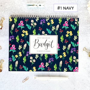 May include: A navy blue 12-month budget planner with a floral pattern of pink, yellow, and purple flowers and green leaves. The word "Budget" is written in cursive on the cover, with the text "Daisy and Decor" above it.