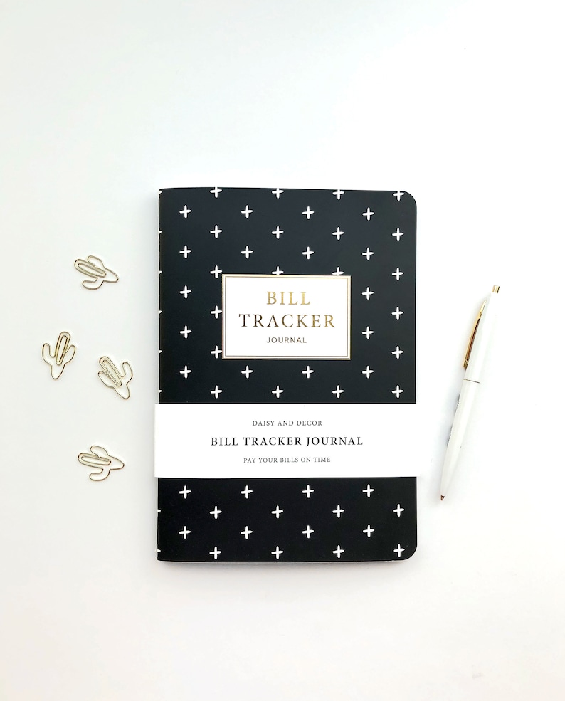May include: A black journal with a white plus sign pattern, labelled "BILL TRACKER JOURNAL". The journal has a white band with the text "PAY YOUR BILLS ON TIME". A white pen and three gold cactus paper clips are also visible.
