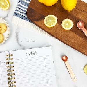 Op de afbeelding: Een wit receptenboek met een gouden spiraalbinding. Het boek heeft een titelpagina met de tekst "Contents" en "Recipe". Het boek ligt op een wit marmeren oppervlak met citroenen, een houten snijplank en maatschepjes.
