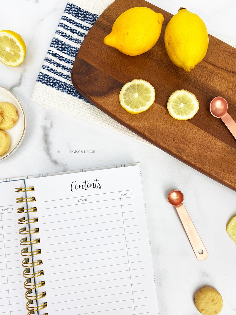 Op de afbeelding: Een wit receptenboek met een gouden spiraalbinding. Het boek heeft een titelpagina met de tekst "Contents" en "Recipe". Het boek ligt op een wit marmeren oppervlak met citroenen, een houten snijplank en maatschepjes.