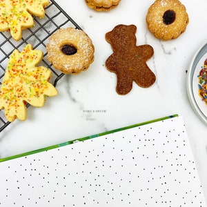 Puede incluir: Una variedad de galletas en una superficie blanca. Hay dos galletas con forma de hoja con chispas de colores, dos galletas redondas con un agujero en el centro y un remolino de mermelada, y una galleta con forma de oso marr&oacute;n. Las galletas est&aacute;n en una rejilla de alambre y un plato blanco con chispas de colores.