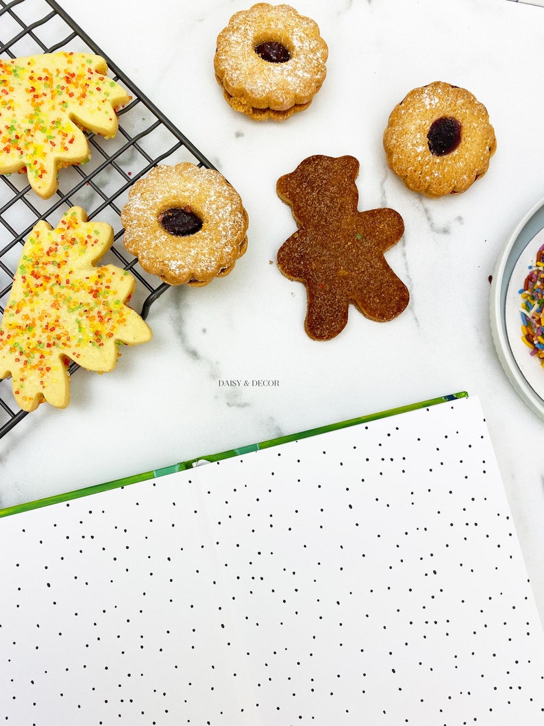 Puede incluir: Una variedad de galletas en una superficie blanca. Hay dos galletas con forma de hoja con chispas de colores, dos galletas redondas con un agujero en el centro y un remolino de mermelada, y una galleta con forma de oso marr&oacute;n. Las galletas est&aacute;n en una rejilla de alambre y un plato blanco con chispas de colores.