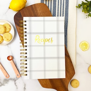 Op de afbeelding: Een wit spiraalgebonden receptenboek met een grijs-wit geruit patroon en het woord "Recepten" in gouden letters.