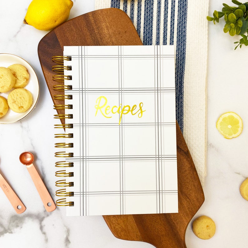 Op de afbeelding: Een wit spiraalgebonden receptenboek met een grijs-wit geruit patroon en het woord "Recepten" in gouden letters.