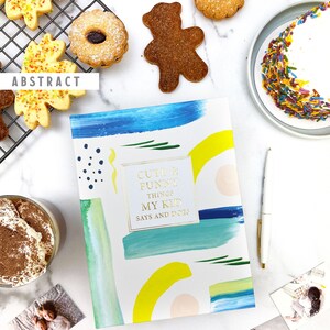 Puede incluir: Un diario azul, amarillo y blanco con el texto "Cute & Funny Things My Kid Says and Does" en letras doradas. El diario est&aacute; sobre una superficie blanca con una variedad de galletas, un plato de chispas y fotos de ni&ntilde;os.
