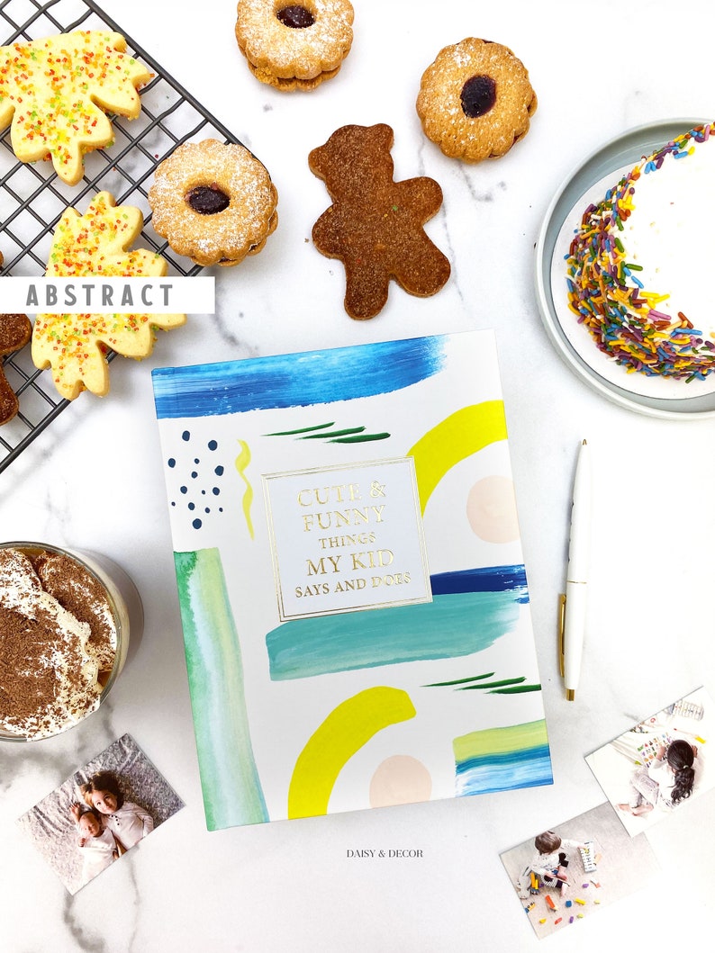 Puede incluir: Un diario azul, amarillo y blanco con el texto "Cute & Funny Things My Kid Says and Does" en letras doradas. El diario est&aacute; sobre una superficie blanca con una variedad de galletas, un plato de chispas y fotos de ni&ntilde;os.