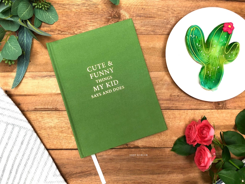 May include: A green journal with the text "Cute & Funny Things My Kid Says and Does" on the cover. The journal is on a wooden surface with a green cactus cookie, a bouquet of pink roses, and a white and gray striped towel.