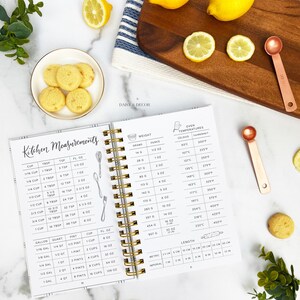 Op de afbeelding: Een wit en grijs spiraalnotitieblok met gouden ringen. Het notitieblok heeft een zwart-witte illustratie van keukenmeetgereedschap en een tabel met keukenmaten in ounces, kopjes, pints, quarts, gallons, eetlepels, theelepels en vloeibare ounces. Het notitieblok bevat ook een tabel met oventemperaturen in Celsius en Fahrenheit. Het notitieblok is open op de pagina met de tabel van de keukenmaten. Het notitieblok ligt op een wit oppervlak met citroenen en een kleine maatschep.