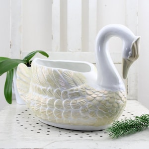 Puede incluir: Macetero de cerámica blanca en forma de cisne con un glaseado amarillo iridiscente. El macetero tiene forma de cisne con la cabeza girada hacia un lado. El cuello del cisne es largo y curvo, y su pico es puntiagudo. El macetero tiene una boca ancha y abierta que es perfecta para contener plantas.