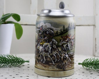Könnte beinhalten: Ein antiker Bierkrug aus Keramik mit einem Zinnverschluss. Der Krug zeigt eine farbenfrohe Illustration einer von Pferden gezogenen Kutsche, die durch einen Wald fährt.