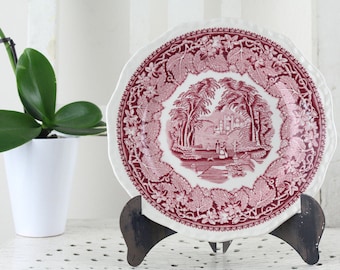 May include: A decorative plate with a red and white design featuring a landscape scene. The plate is displayed on a black metal stand. A white potted plant with green leaves is to the left of the plate.