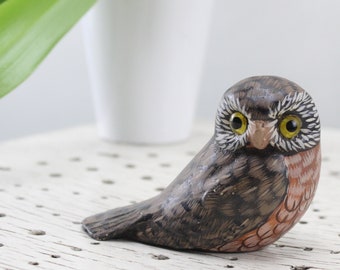May include: A small, hand-carved wooden owl figurine with brown, gray, and orange paint. The owl has large, yellow eyes and a detailed beak.