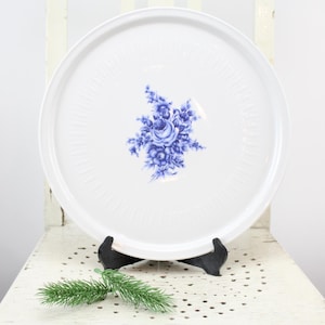 Könnte beinhalten: Ein weißer, runder Servierteller mit einem blauen Blumendesign in der Mitte. Der Teller wird auf einem weißen Stuhl mit einem schwarzen Ständer und einem Zweig Grün präsentiert.