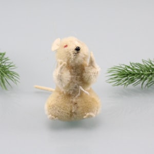 Puede incluir: Un ratón de peluche blanco con ojos rojos y nariz negra. El ratón está de pie sobre sus patas traseras con las patas delanteras levantadas. El ratón está sobre un fondo azul claro con ramas de pino verdes a cada lado.