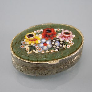 Könnte beinhalten: Eine kleine ovale Schmuckschatulle mit einem floralen Mosaikdesign. Das Mosaik zeigt eine Vielzahl von bunten Blumen, darunter rote, gelbe, rosa und blaue, auf grünem Hintergrund. Die Schachtel ist aus Metall und hat eine silberne Oberfläche.
