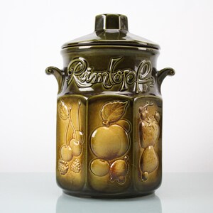 May include: A green ceramic Rumtopf jar with a lid and handles. The jar features embossed fruit designs, including cherries, apples, and pears. The word "Rumtopf" is written across the top in a decorative font. This jar is likely used for preserving fruit.