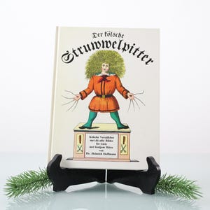Könnte beinhalten: Ein Vintage-Buch mit dem Titel "Der töltsche Struwwelpitter" wird auf einem schwarzen Ständer mit einem Zweig Grünzeug präsentiert. Das Buchcover zeigt eine farbenfrohe Illustration einer Figur mit wildem grünem Haar, einem orangefarbenen Mantel und grünen Hosen.