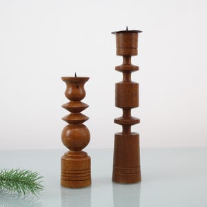 Puede incluir: Dos candelabros de madera de diferentes alturas. El más corto tiene una base redondeada y una sección media bulbosa, mientras que el más alto es más columnar. Ambos son de color marrón cálido y tienen un diseño vintage simple.