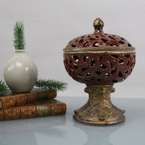 Könnte beinhalten: Ein roter Keramik-Räucherofen mit dekorativem goldenen Rand und einem komplizierten Ausschnittmuster. Der Ofen steht auf einem goldenen Sockel mit einem Blattdesign.