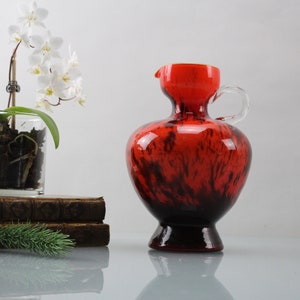 Puede incluir: Una jarra de vidrio roja y negra con un asa transparente. La jarra tiene un diseño moteado, marmoleado.