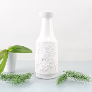以下が含まれることがあります： 葉のレリーフ模様が施された白い陶器の花瓶。花瓶は背が高く、細身で、丸みを帯びた底とわずかに広がった口をしています。