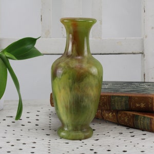 May include: A green and brown glazed ceramic vase with a narrow neck and a rounded base. The vase is sitting on a white surface with a white chair behind it.