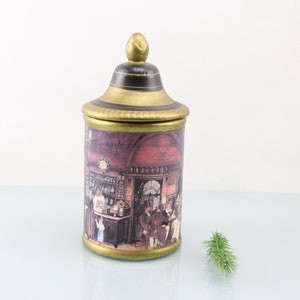 May include: A ceramic jar with a gold lid and a scene of a vintage apothecary shop printed on the side. The jar is on a white surface.