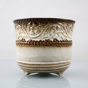 May include: A white ceramic planter with a brown floral design around the top rim. The planter has three small feet and a rounded base.