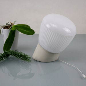 May include: A white ceramic table lamp with a ribbed shade and a white cord. The lamp is on a grey surface with a small potted plant in the background.