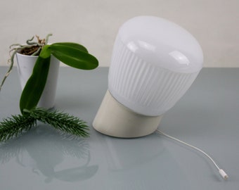 May include: A white ceramic table lamp with a ribbed shade and a white cord. The lamp is on a grey surface with a small potted plant in the background.