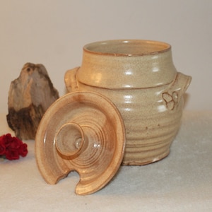 Puede incluir: Un tarro de cerámica beige con tapa. El tarro tiene dos asas y una superficie texturizada. La tapa tiene un pequeño pico.