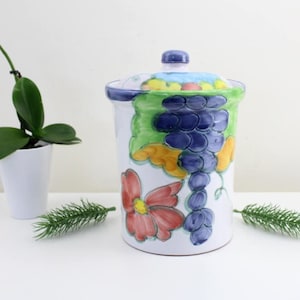 Puede incluir: Jarrón de cerámica blanca con tapa, con un diseño pintado a mano de uvas, hojas y flores en tonos azules, verdes, amarillos y rojos.