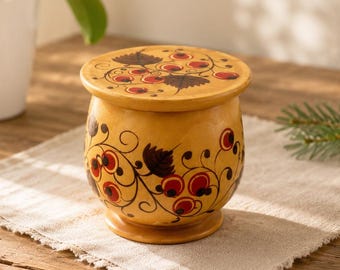 May include: A small, round, yellow wooden container with a lid, featuring hand-painted black swirling vine patterns and clusters of red berries on both the lid and the main body. The container has a smooth, polished finish and a pedestal base.