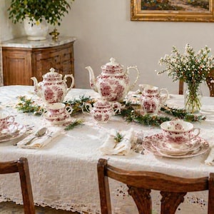 Könnte beinhalten: Ein formeller Esstisch, der für Tee gedeckt ist. Der Tisch ist mit einer weißen Spitzentischdecke bedeckt und mit einem floralen Teeservice gedeckt, einschließlich Tassen, Untertassen und einer Teekanne. Der Tisch ist mit Grün und Blumen dekoriert.