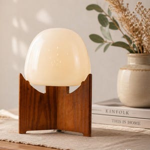 May include: A retro-style table lamp featuring a cream-coloured, rounded glass shade, set on a dark wooden base with a distinctive design. The lamp is placed on a wooden table with a beige runner. A vase with dried flowers is also visible.