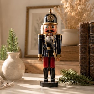 May include: A wooden nutcracker figurine wearing a black helmet with a gold emblem, a dark blue uniform with gold trim, and red trousers. The figure has a painted face with a moustache and holds a thin, long staff in its right hand.