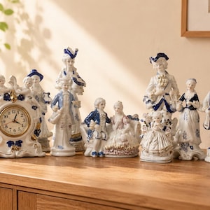 May include: A collection of white porcelain figurines with blue and gold accents, including a mantle clock with a round face and Roman numerals, alongside several standing and seated figures dressed in historical attire, all arranged on a wooden surface.