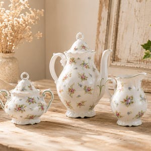 Puede incluir: Un juego de té de porcelana blanca con un delicado estampado floral de pequeñas flores rosas, azules y amarillas con hojas verdes. El conjunto incluye una tetera con tapa, un azucarero con tapa y dos asas, y una jarra de leche con bordes festoneados.